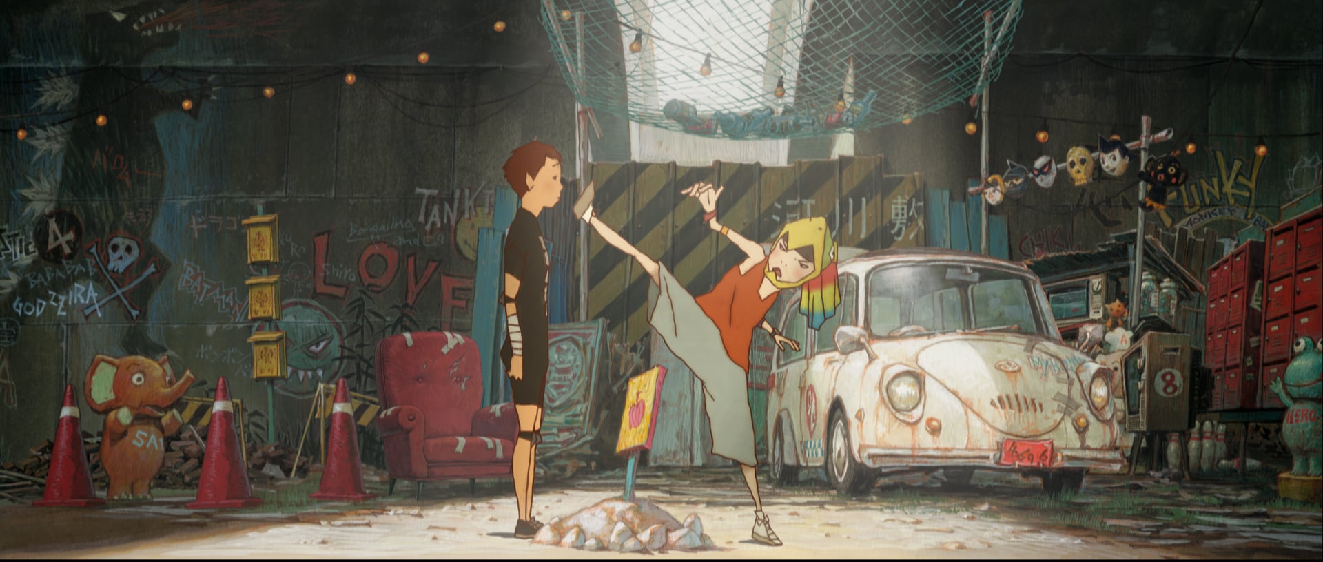 A girl kicks high near a boy in a cluttered garage with graffiti, an old white car, a red chair, and various objects scattered around.