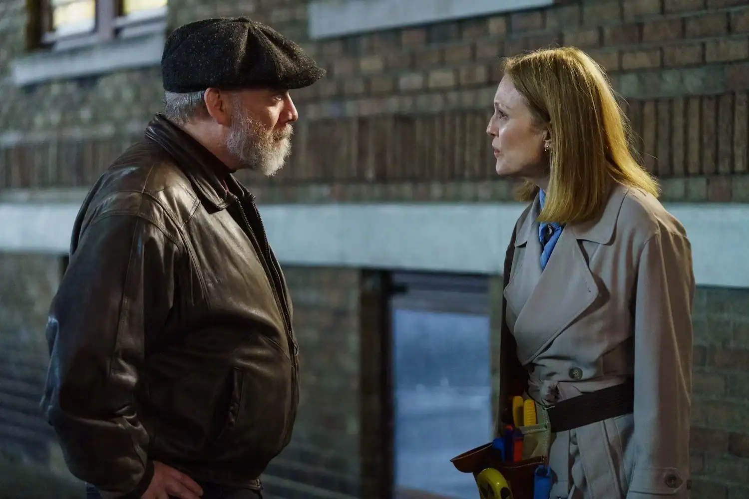 A man in a leather jacket and cap faces a woman in a trench coat with tools in her belt, standing outside near a brick wall, having a serious conversation.