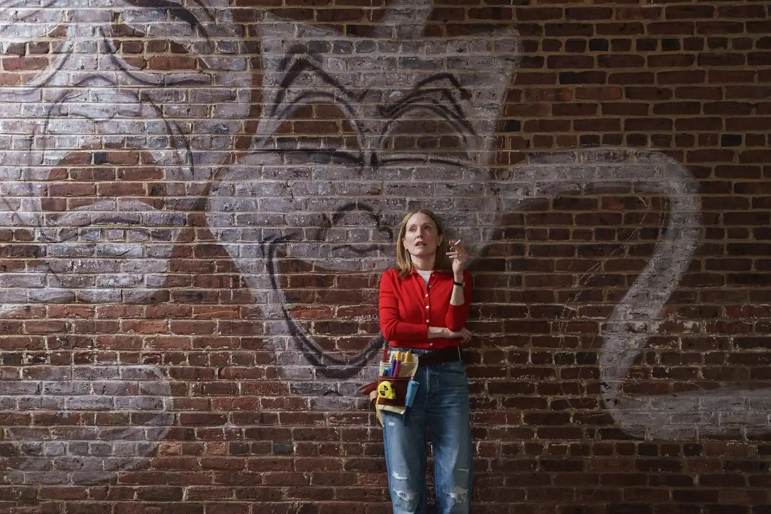 A woman in a red sweater and ripped jeans stands against a graffiti-covered brick wall, holding a cigarette, with a large cartoon face behind her.