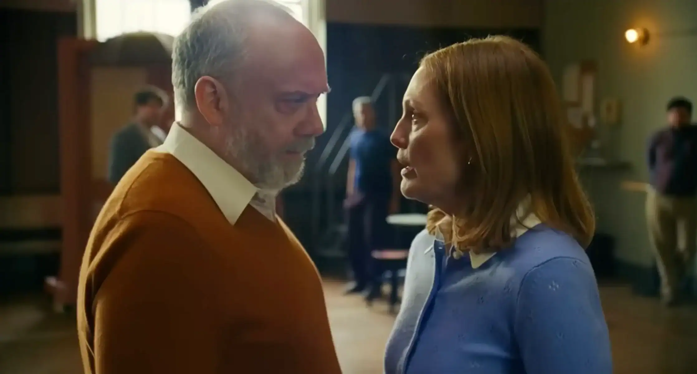 A man and woman stand close together indoors, looking at each other seriously, with other people blurred in the background.