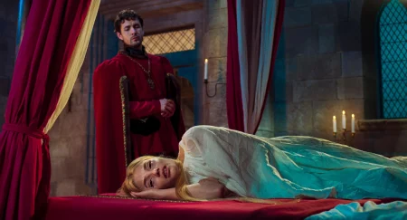 A woman in a white dress lies on a red bed, appearing distressed, while a man in a red robe stands behind her in a dimly lit stone room with candlelight.
