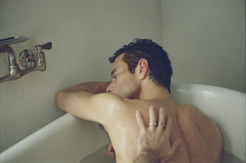 A man with wet hair rests his head on the side of a bathtub while another person’s hand gently touches his back.