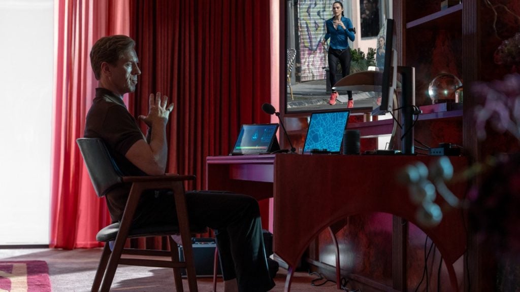 Person sitting at a desk with laptops, watching another person jogging on a large screen in a room with red curtains.