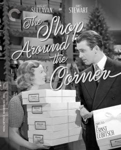 A woman and a man in a suit stand facing each other in a shop, holding boxes, with “The Shop Around the Corner” movie title text over the image.