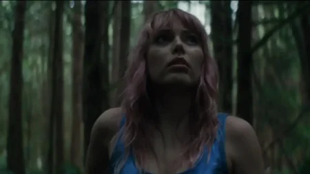 A person with pink hair and a blue top looks upward in a dim, dense forest with tall trees in the background.