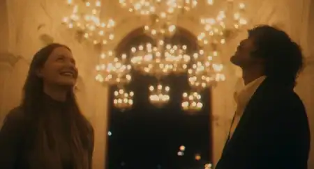 Two people stand indoors under warm, glowing chandeliers, looking up and smiling, with blurred lights in the background.