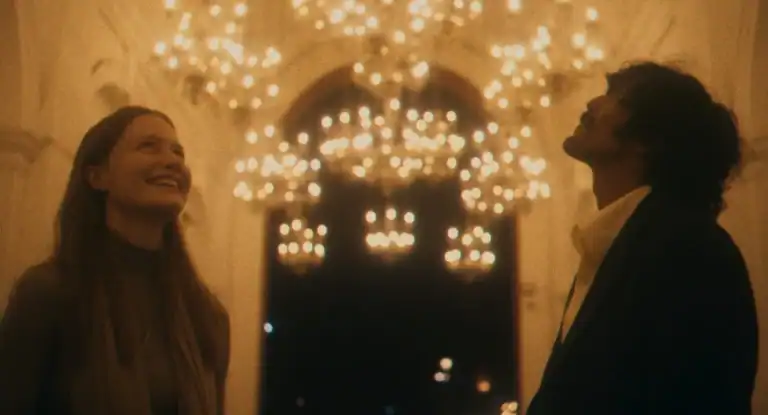 ‘The Cycle Of Love’ Review – Journeys End When Lovers Meet Two people stand indoors under warm, glowing chandeliers, looking up and smiling, with blurred lights in the background.