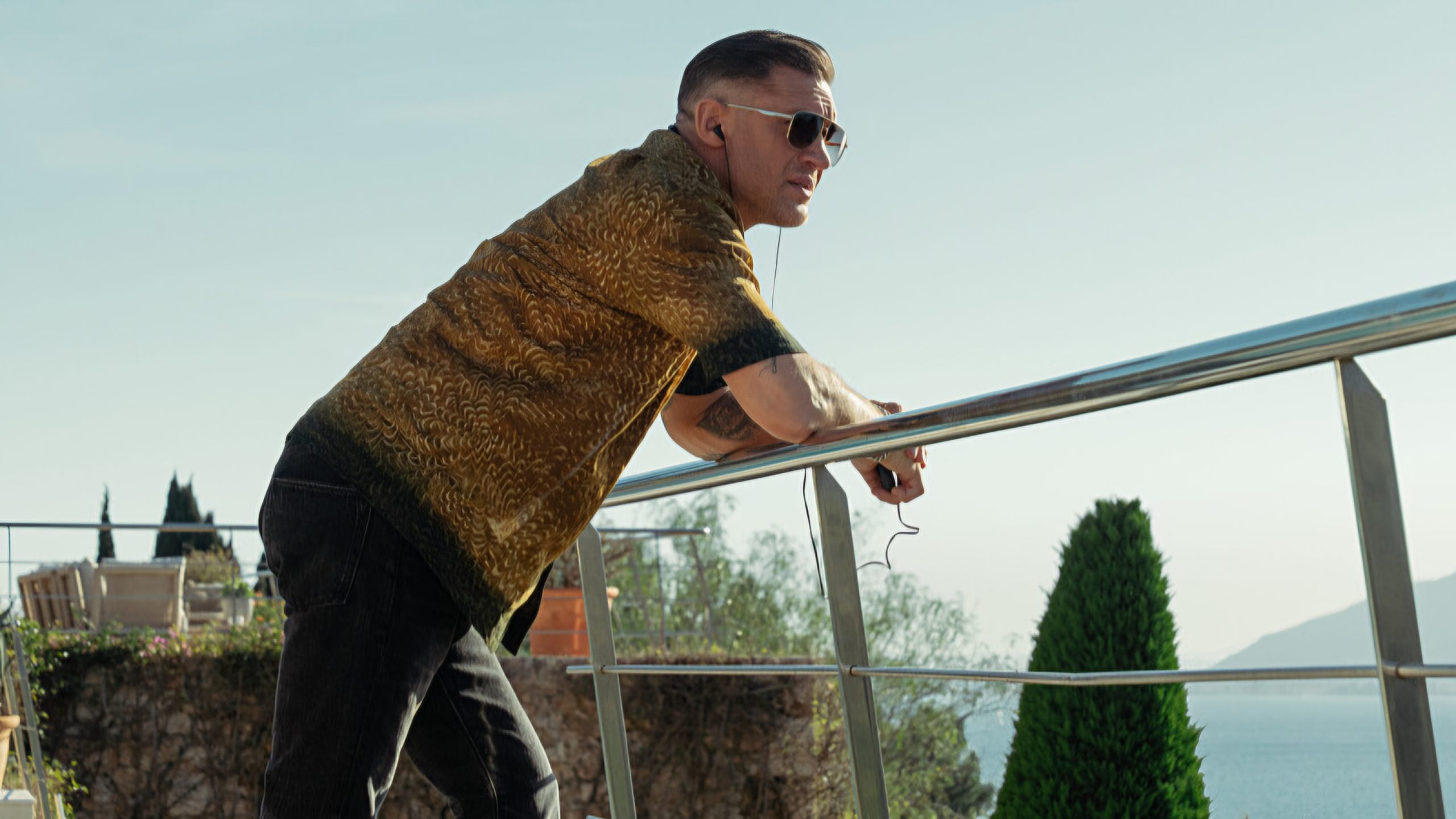 A man in sunglasses and a patterned shirt leans on a metal railing outdoors, looking into the distance, with greenery and clear sky in the background.