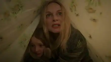 A woman and a young girl huddle together under a floral sheet, both appearing frightened and looking upward.