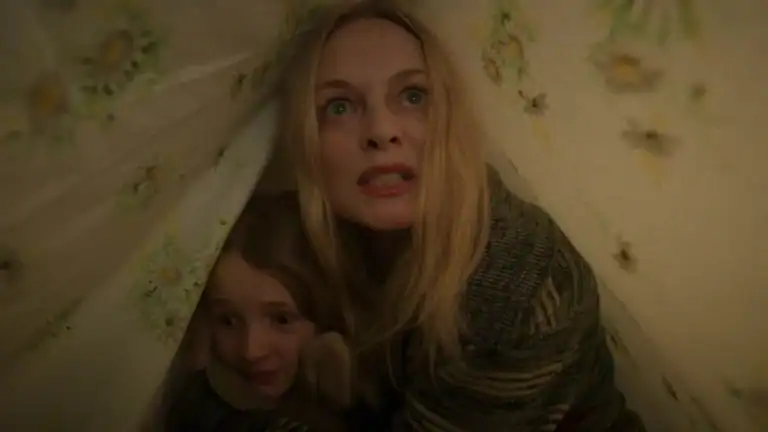 A woman and a young girl huddle together under a floral sheet, both appearing frightened and looking upward.
