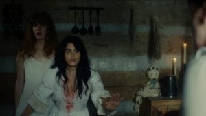 Two women in white nightgowns, one with blood on her chest, stand in a dimly lit rustic room with candles, kitchen utensils, and a cloth doll on a cabinet.