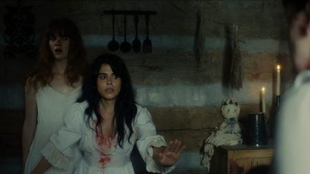 Two women in white nightgowns, one with blood on her chest, stand in a dimly lit rustic room with candles, kitchen utensils, and a cloth doll on a cabinet.
