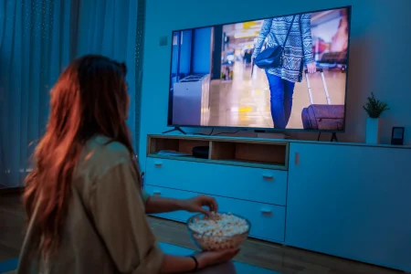 A person sits on the floor, eating popcorn and watching television, which displays someone walking in an airport with luggage.