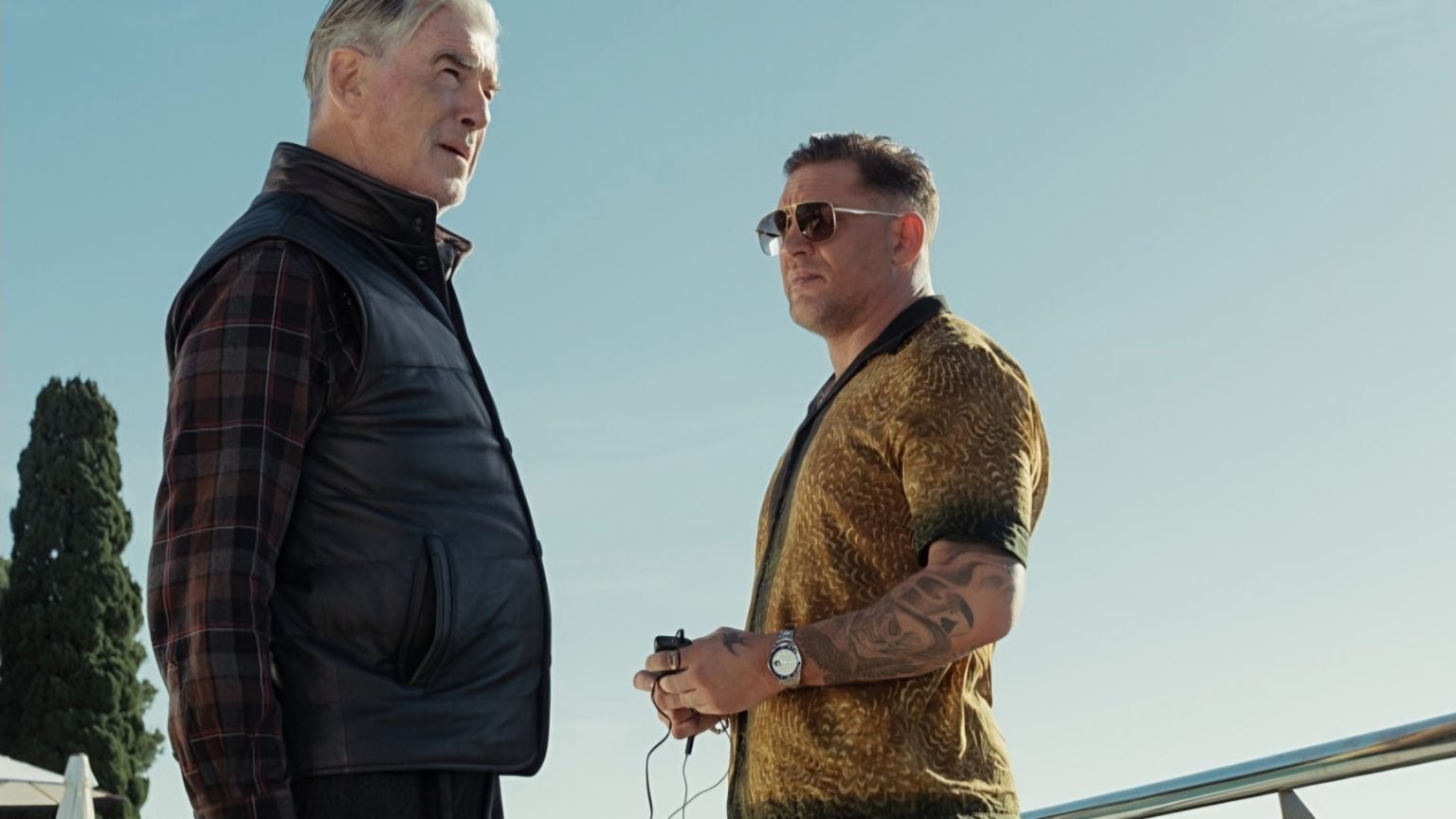 Two men stand outdoors under a clear sky, one wearing a black vest and plaid shirt, the other in a gold shirt with tattoos and sunglasses, holding an object in his hands.