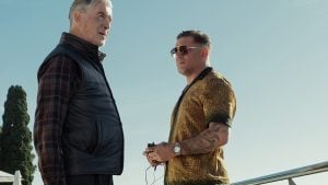 Two men stand outdoors under a clear sky, one wearing a black vest and plaid shirt, the other in a gold shirt with tattoos and sunglasses, holding an object in his hands.