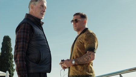 Two men stand outdoors under a clear sky, one wearing a black vest and plaid shirt, the other in a gold shirt with tattoos and sunglasses, holding an object in his hands.