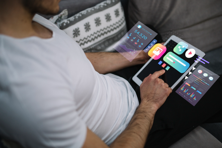 A person in a white shirt uses a tablet on their lap, interacting with various colorful digital charts and icons displayed as augmented reality graphics above the device.