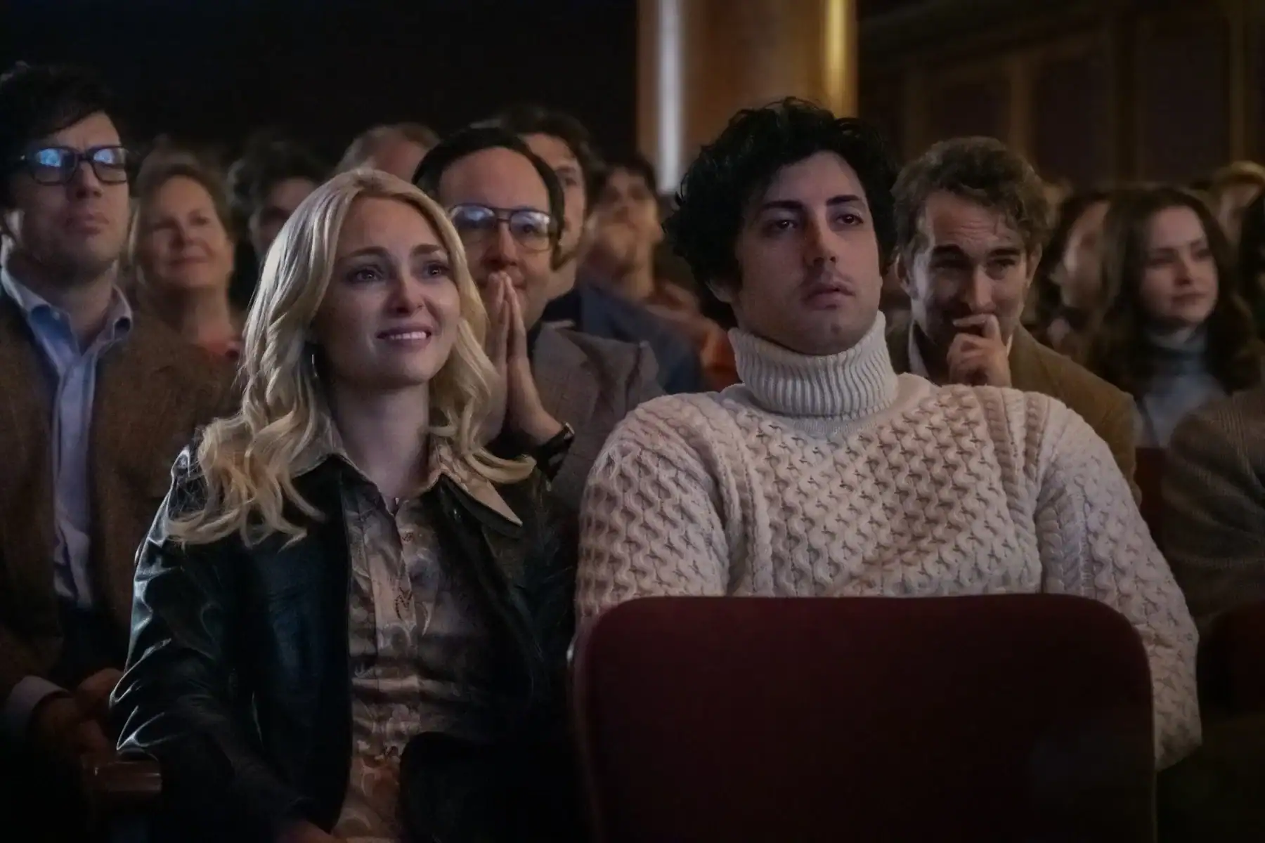 A group of people sit in theater seats, watching a performance. The audience members have neutral or slightly engaged expressions.