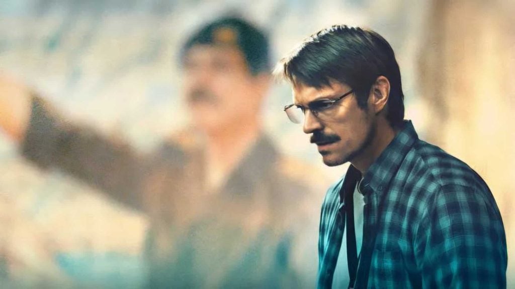 A man with glasses and a mustache, wearing a plaid shirt and tie, stands in focus with a blurred figure in the background.