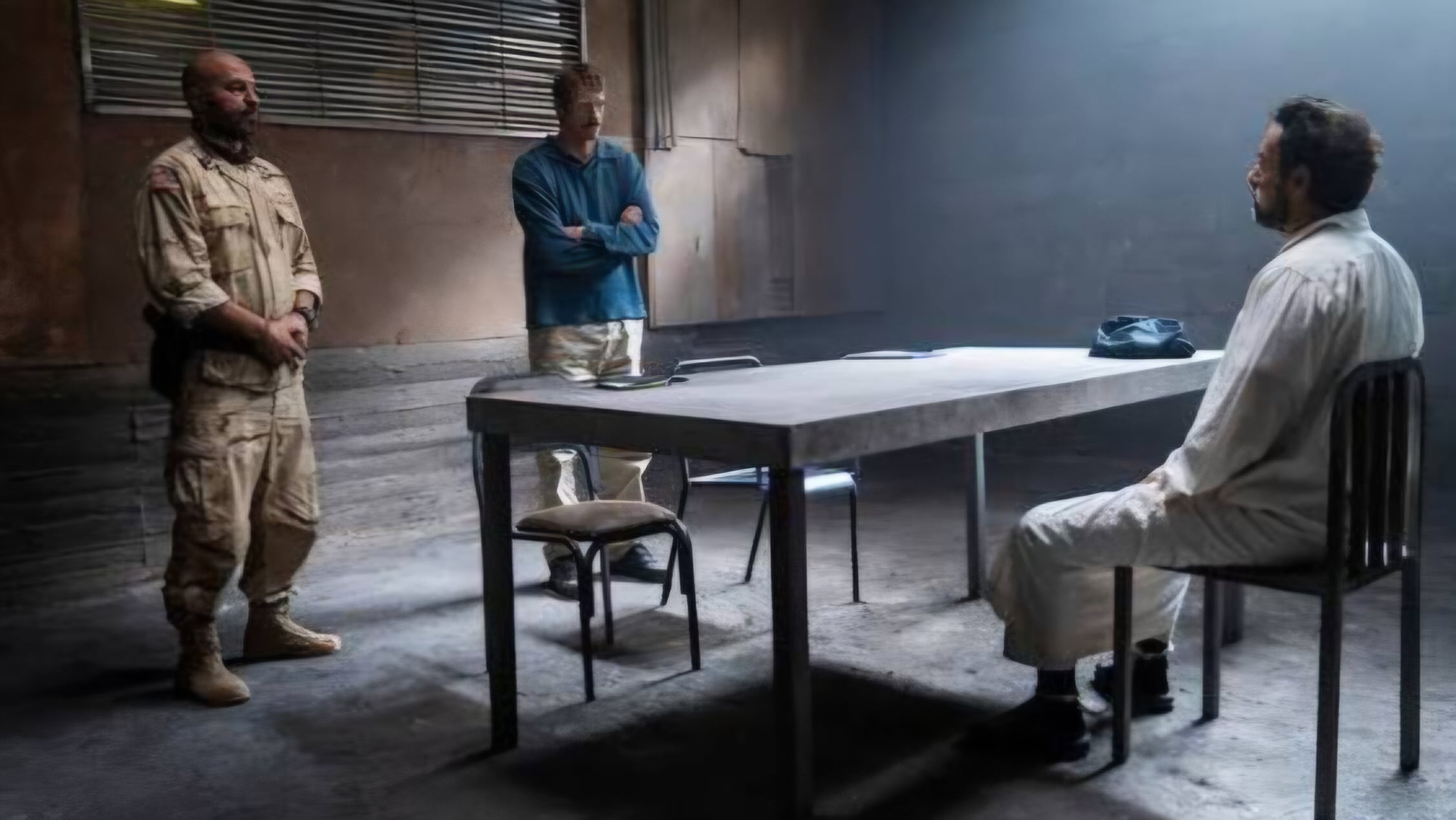 Two men stand while another man, in white clothing, sits at a table in a dimly lit interrogation room with concrete walls and minimal furniture.