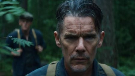 A man with slicked hair and a serious expression walks through a forest, with another man in the background also wearing a backpack.
