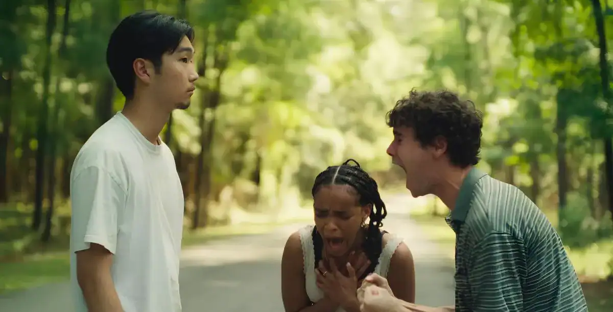 Three people stand on a wooded road; one looks calm, another yells, and the third crouches, appearing distressed.