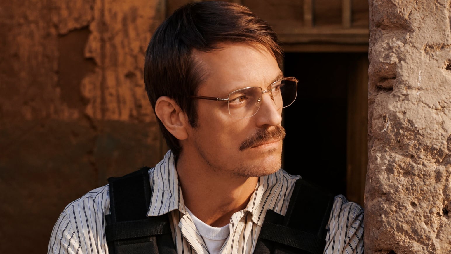 A man with glasses and a mustache, wearing a striped shirt and black vest, looks to the side against an outdoor rustic wall background.