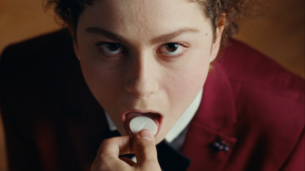 A person in a burgundy jacket puts a white tablet on their tongue, looking upward with a neutral expression.