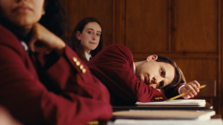 A student in a red blazer rests his head on a desk during class, looking tired, while other students sit nearby.