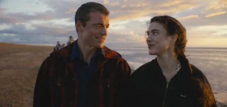 A man and woman walk side by side on a beach at sunset, looking at each other and smiling, with a few people in the background.