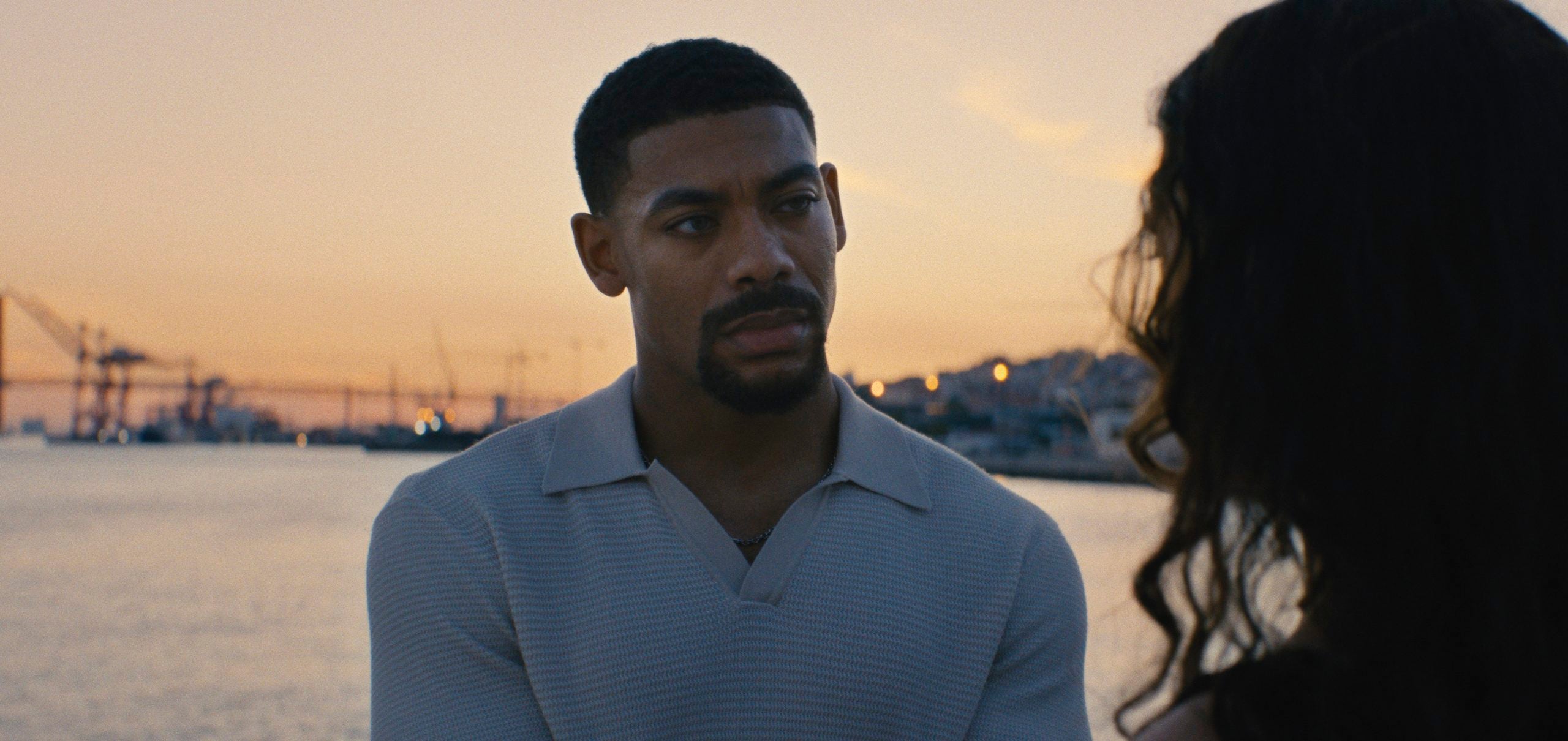 A man in a light sweater stands by the water at sunset, looking at a woman whose back is to the camera. Industrial cranes are visible in the background.
