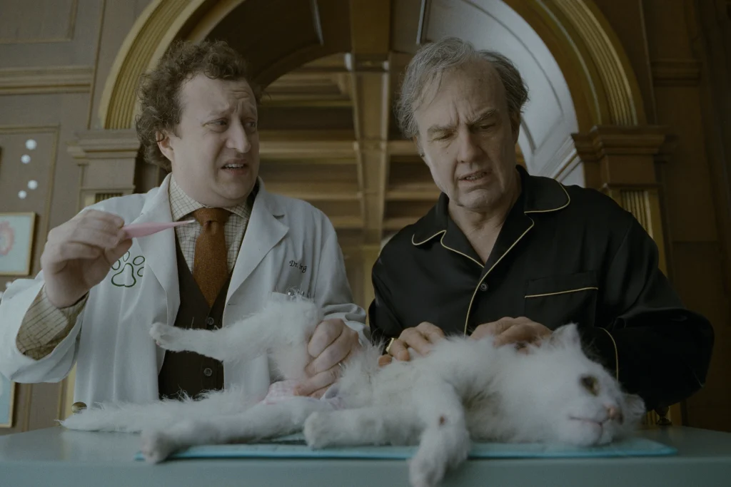 Two men stand over a white cat on an exam table; one wears a lab coat holding tweezers, while the other in pajamas examines the cat.