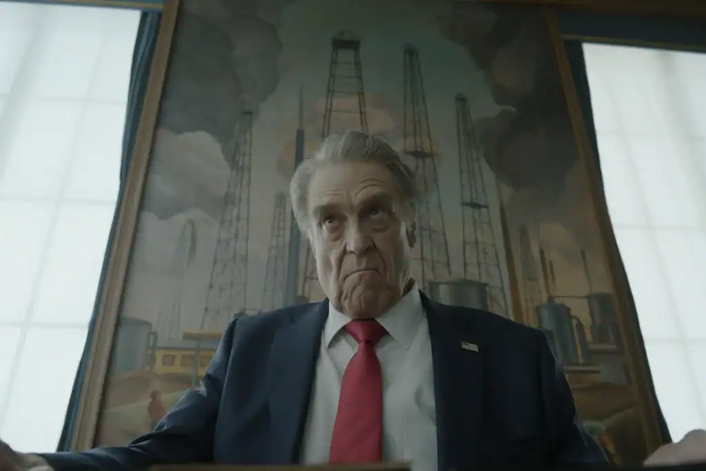 An older man in a suit and red tie sits in front of a large painting of oil rigs, looking upward with a serious expression.