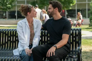 A woman in a white lab coat and a man in a black T-shirt sit closely on a park bench, facing each other and smiling, with trees and people in the background.