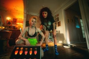 Two people in shorts and socks examine a case of glowing vials on the floor in a dimly lit, cluttered living room, while a third person sits in the background.