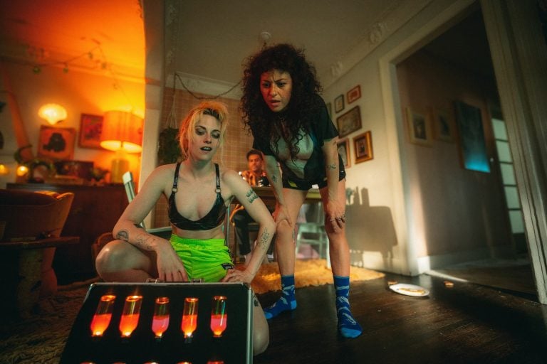 Two people in shorts and socks examine a case of glowing vials on the floor in a dimly lit, cluttered living room, while a third person sits in the background.