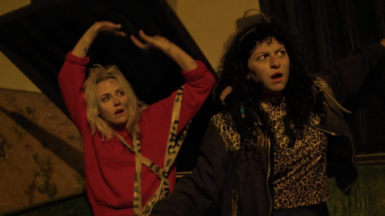Two women stand outside at night with surprised expressions; one in a red sweatshirt raises her arms, the other wears a leopard-print top and spreads her arms wide.