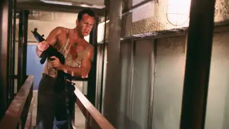 A man in a bloodstained tank top holds a submachine gun while standing in a narrow corridor.