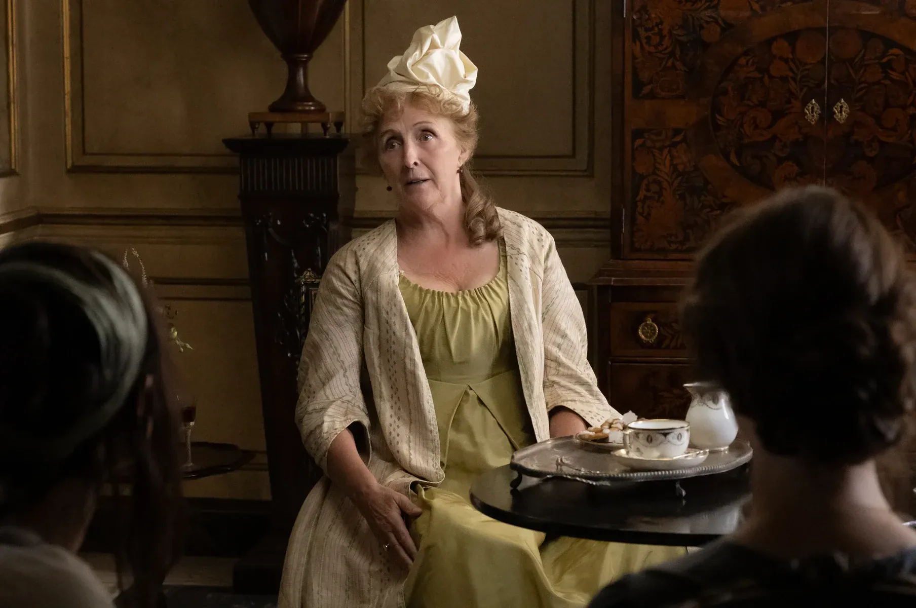 An older woman in a period-style gown and headpiece sits at a small table with tea service, facing two people in the foreground.