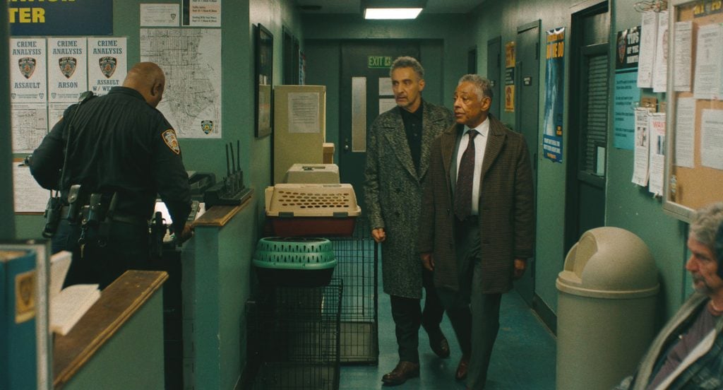 A police officer in a precinct station faces a counter while two men in overcoats walk down the hallway behind him.
