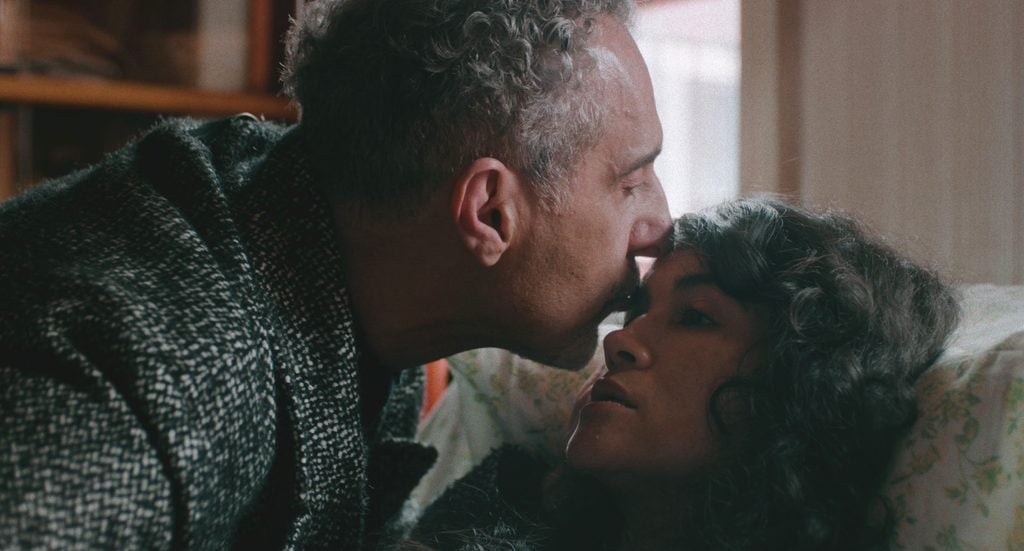An older man kisses a younger woman on the forehead as she lies in bed.
