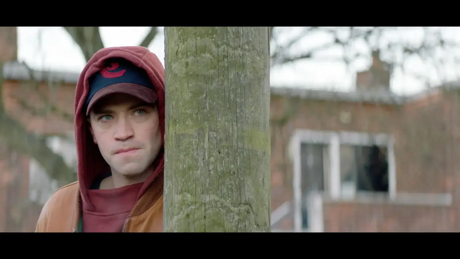 A man in a hooded jacket and baseball cap peeks from behind a wooden pole.
