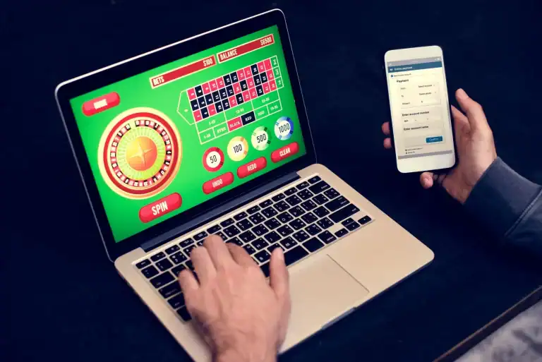 Geek Culture Meets Online Gaming: A Look at JooCasino A person using a laptop displaying an online roulette game while holding a smartphone open to an online payment screen.