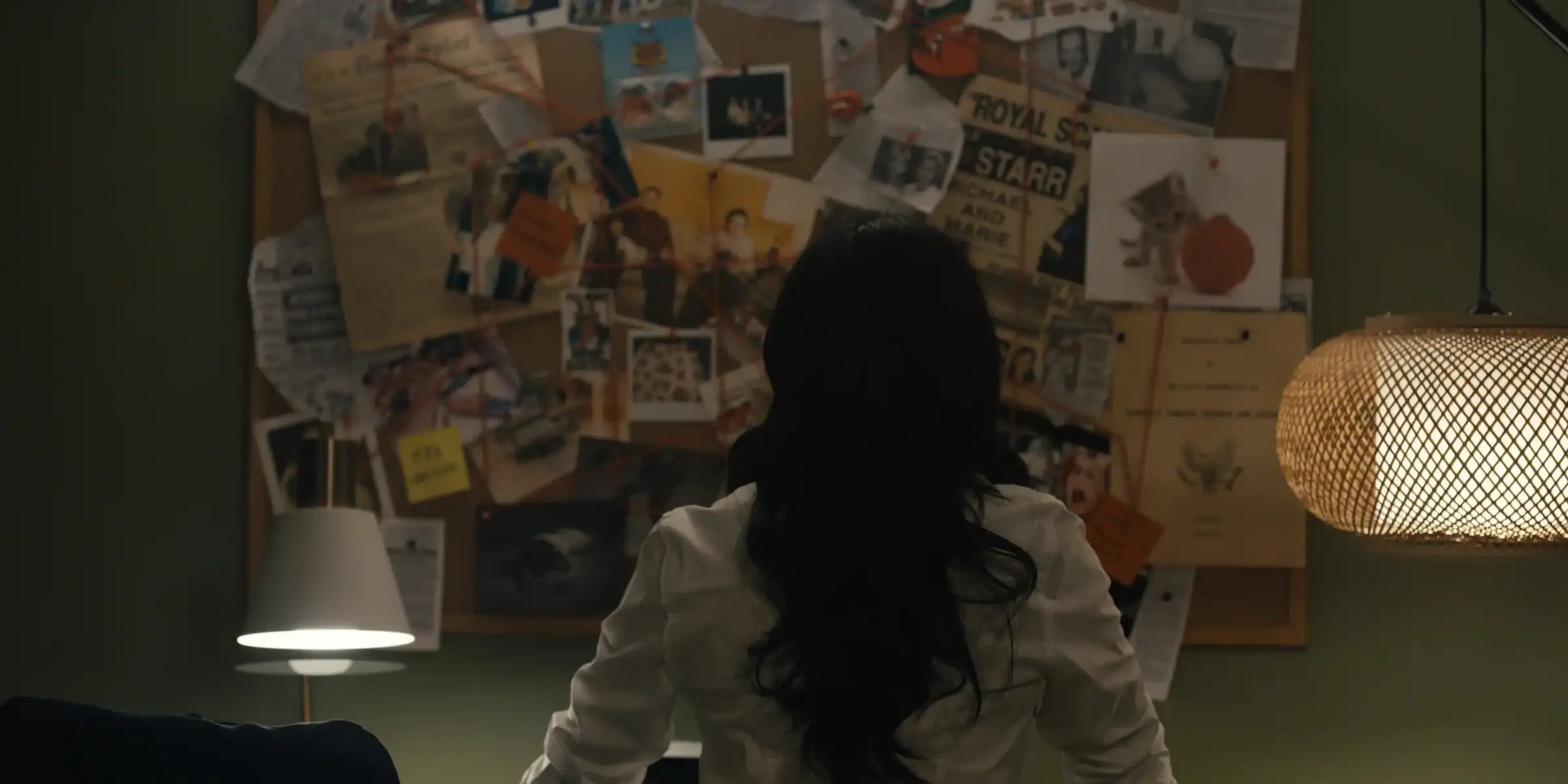 A person viewed from behind faces a corkboard covered in photographs, documents, and red string.