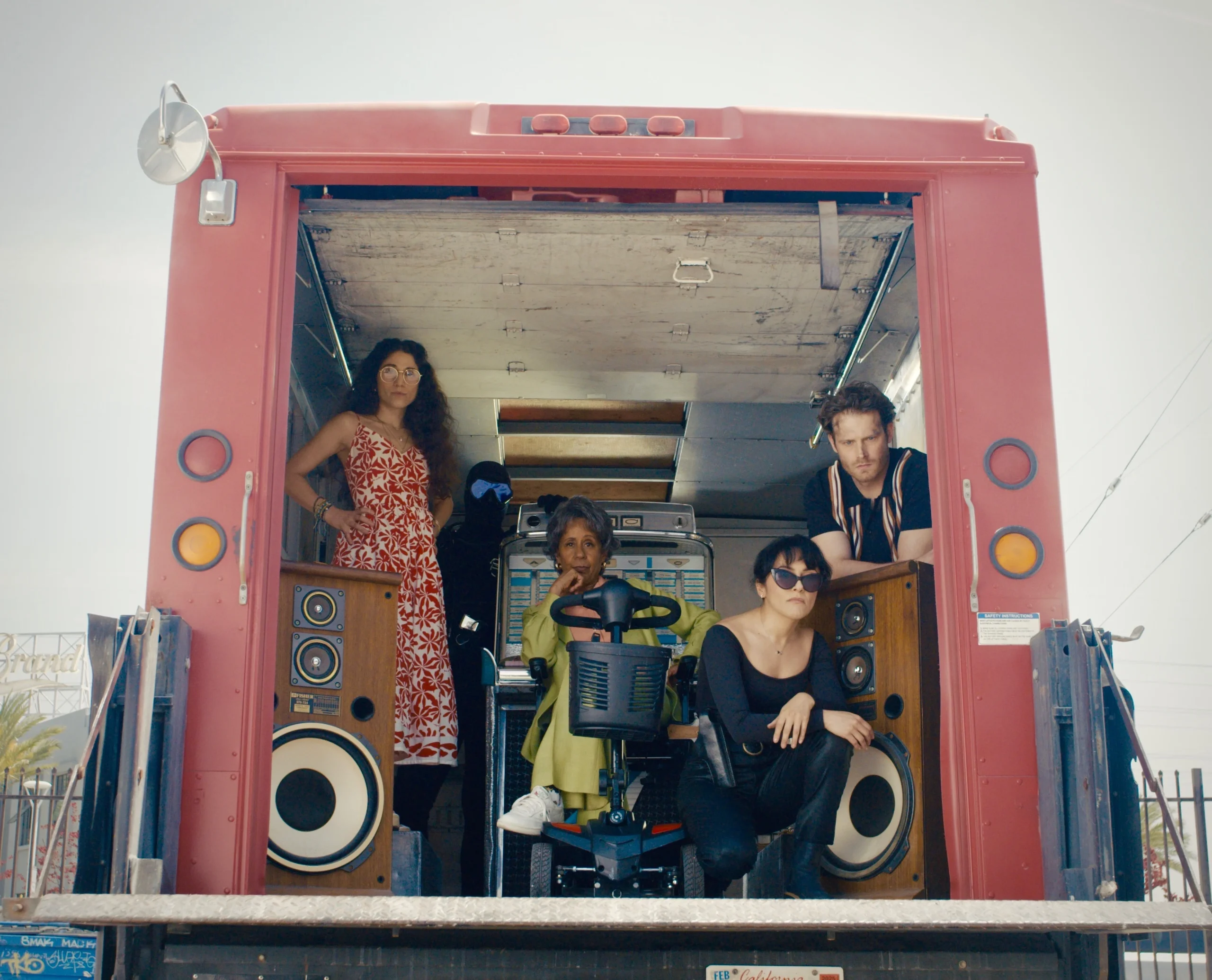 Five people pose inside the back of a red truck, with two large speakers on either side and a mobility scooter in the center.