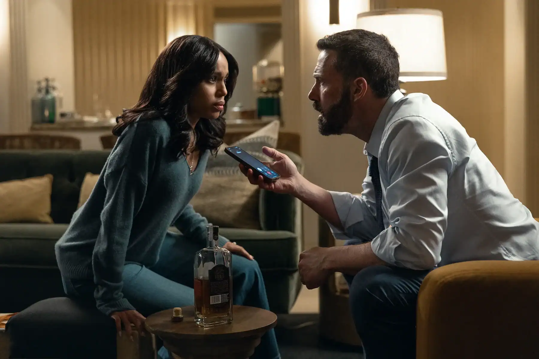 A woman sits on a couch looking at a man kneeling before her, who holds up a smartphone during a call. A bottle of whiskey sits on a small table between them.