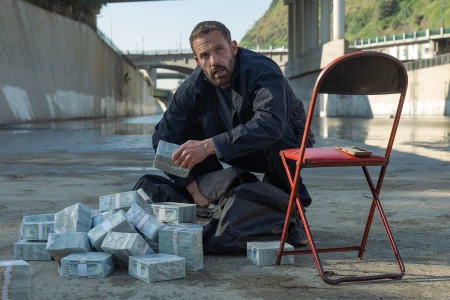 A man crouches in a concrete drainage channel next to a red folding chair and several stacks of bundled hundred-dollar bills.