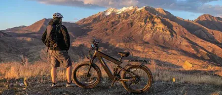 A cyclist with a backpack stands with an electric mountain bike overlooking a vast mountain range at sunset.