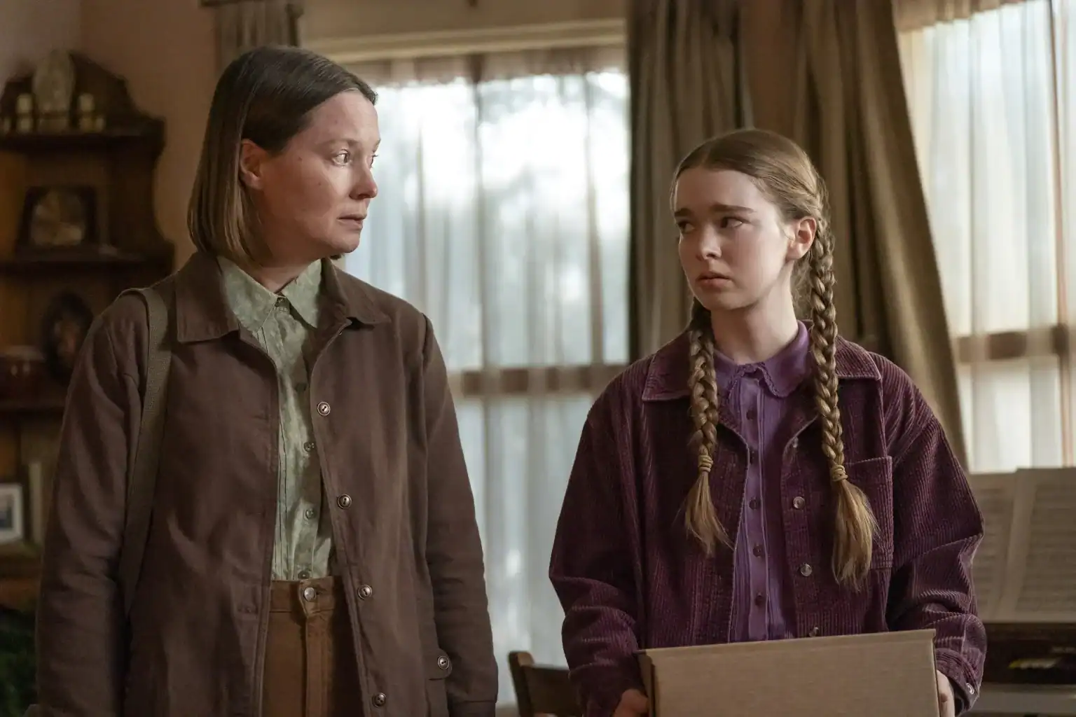 An adult woman and a young girl with braided hair look toward each other while holding a cardboard box.
