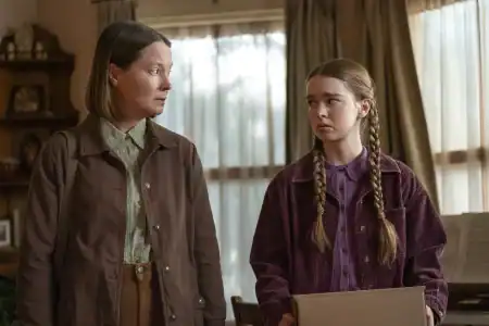 An adult woman and a young girl with braided hair look toward each other while holding a cardboard box.